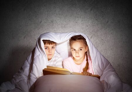 Little girl and boy looking at each other under blanket. Children reading magic stories in bed before going to sleep. Young sister and brother in pajamas together on background of grey wall.の写真素材