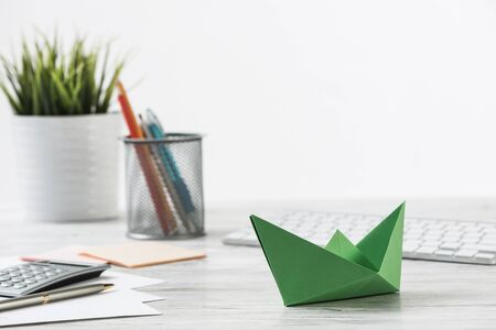 Wooden office desk with green origami boat. Individual investment and money saving. Flat lay office workplace with plant in pot, computer keyboard and calculator. Green technologies and innovationsの写真素材