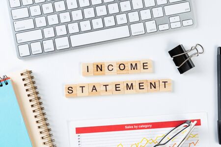 Income statement concept with letters on wooden cubes. Still life of office workplace with supplies. Flat lay white surface with computer keyboard and analytic report. Financial success and investmentの写真素材
