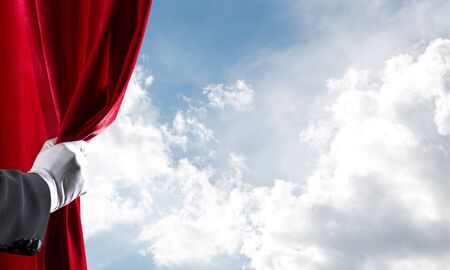 Human hand in glove opens red velvet curtain on blue sky backgroundの写真素材
