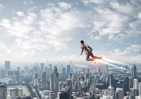 Businessman in aviator hat flying in blue sky as superhero. Business person as superhero with jet flame flying above modern metropolis. Goal achievement concept. Career ambition and growth.の写真素材