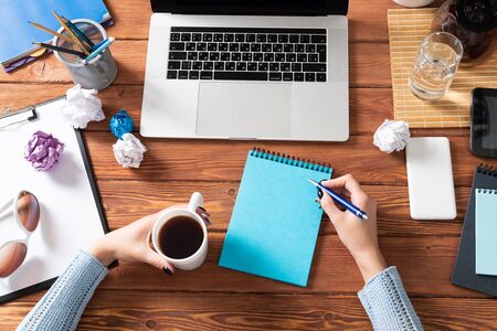 Woman writing in notebook with pen at desk. Online business education. Flat lay office workplace with female hands and laptop. Digital technology and coworking space. Female journalist taking notesの写真素材