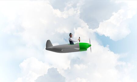 Man in aviator hat with goggles driving propeller plane. Funny man having fun in small airplane. Blue cloudy sky with fluffy clouds. Businessman sitting in paper plane and holding steering wheel.の写真素材