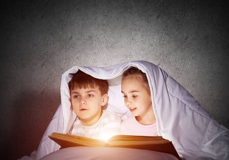 Smiling kids reading magic fairy tales in bed. Happy children with flashlight hiding under blanket. Little friends having fun at bedtime. Young boy and girl in pajamas spend time together in bedroom.の写真素材