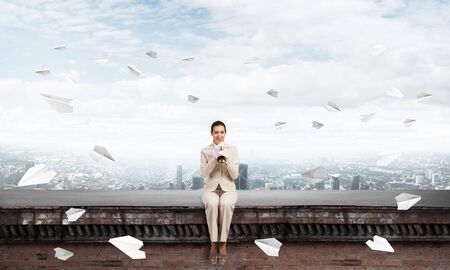 Beautiful young woman playing trumpet on roof. Brave girl in white business suit with music brass instrument sitting on edge of roof. Mixed media business concept. Flying paper planes in blue skyの写真素材