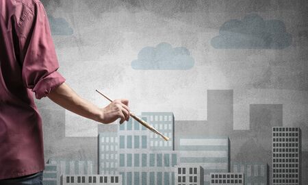 Close up artist hand holding paintbrush. Painter in shirt standing on background colorful picture of city. Modern cityscape with high skyscrapers artwork. Creative hobby and artistic occupation.の写真素材
