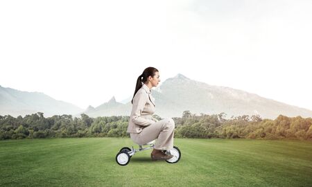 Beautiful young woman riding childrens bicycle on green grass. Businesslady in white business suit...