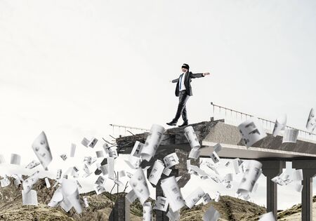 Businessman walking blindfolded among flying documents on concrete bridge with huge gap as symbol of hidden threats and risks. Skyscape and nature view on background. 3D rendering.の写真素材