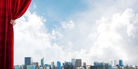 Human hand opens red velvet curtain to modern cityscape and cloudy skyの写真素材