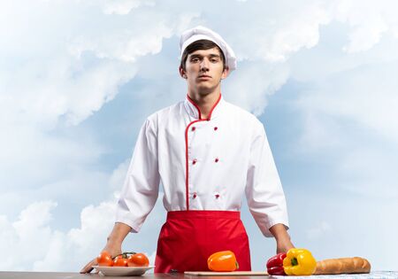 Young male chef standing near cooking table with vegetables. Handsome chef in white hat and red apron on blue sky background. Cooking classes advertising. Restaurant food preparation conceptの写真素材