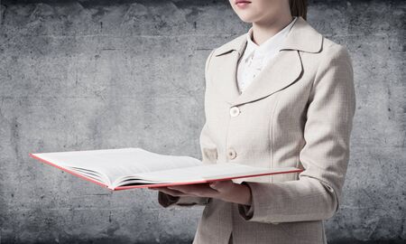 Corporate employee holding open notebook on background of grey wall. Business paperwork and accounting. Closeup open book with red cover in female hands. Elegant young woman in business suit.の写真素材