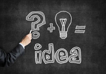 Businessman hand with chalk draws idea generation sketch on chalkboard. Freehand white chalk illustration on blackboard. Question mark and lightbulb symbols. Brainstorming business ideas conceptの写真素材