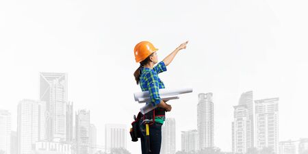 Female engineer in hardhat standing with technical blueprints and pointing upwards. Side view of woman architect in checkered blue shirt inspecting construction. Building project management and audit.の写真素材