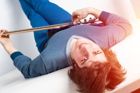 Happy young man lying back on sofa with guitar. Smiling guitarist relaxing on white sofa. Close-up caucasian musician playing electric guitar and looking at camera. Playing musical instrumentsの写真素材