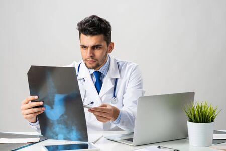 Handsome young doctor looking at x-ray scan. Physician in white coat with stethoscope sitting at...