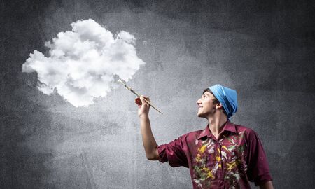 Young artist drawing realistic white cloud. Male painter in dirty shirt and bandana with paintbrush on grey wall background. Creative hobby and artistic occupation. Art classes concept with copy spaceの写真素材