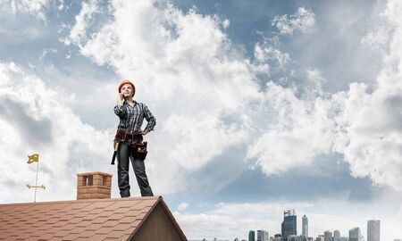 Attractive blonde woman in workwear and hardhat talking on smartphone. Young worker in checkered blue shirt standing on brick roof of building. Professional renovation and roofing construction.の写真素材