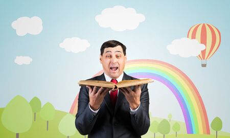 Surprised businessman holding open old book. Startled adult man in business suit and tie standing on background of nature landscape with greenfield and rainbow illustration. Education and knowledges.の写真素材