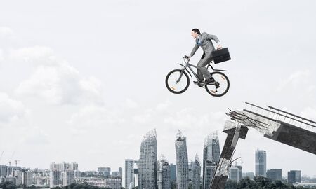 Man on bike jumping from broken bridge. Businessman on bike hurry to work. Corporate employee in business suit with suitcase on cycle flying in air. Cyclist on background modern cityscape and blue skyの写真素材