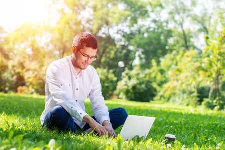 Young businessman sitting on green grass and using laptop computer. Handsome man working with computer in park at sunny summer day. Outdoors nature journey and relaxation. Freelance work concept.の写真素材