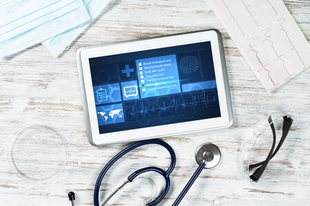 Modern social health insurance program. Tablet computer with healthcare application interface on screen. Top view doctor workplace with stethoscope and cardiogram. Digital healthcare technologies.の写真素材