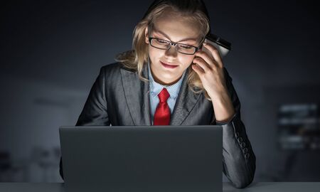 Portrait of concentrated businesswoman in glasses sitting at office desk and working at laptop. Financial consultant in business suit and tie busy at work in evening. Working in office late at night.の写真素材