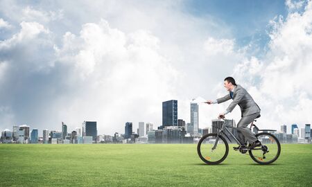 Businessman with paper documents in hand on bike. Paperwork express service. Corporate employee in business suit riding bicycle on green field. Cyclist on background of blue sky and city on horizonの写真素材