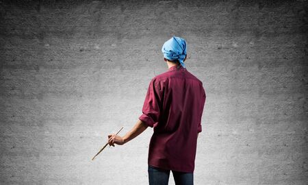 Young male painter artist holding paintbrush. Back view of painter in shirt and bandana standing on gray wall background. Creative hobby and artistic occupation. Art school courses concept.の写真素材