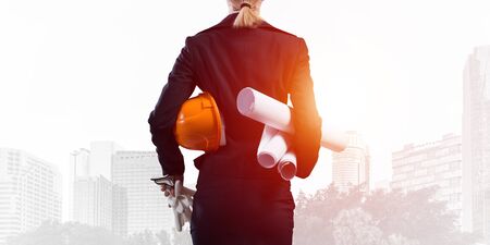 Woman architect standing with construction drawings and hardhat in hands. Back view of contractor on background of modern office buildings. Industrial engineering and construction projects management.の写真素材