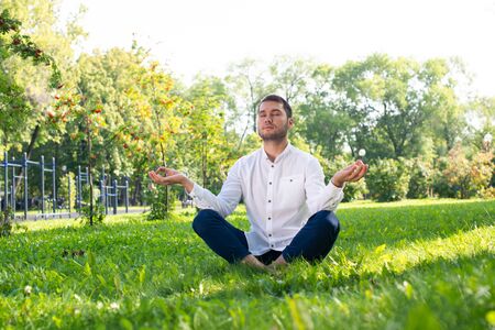 Young man meditates in lotus pose on green grass. Handsome man in casual wear practicing of yoga with closed eyes. Training and meditation outdoor at summer day. Healthy lifestyle and relaxation.の写真素材