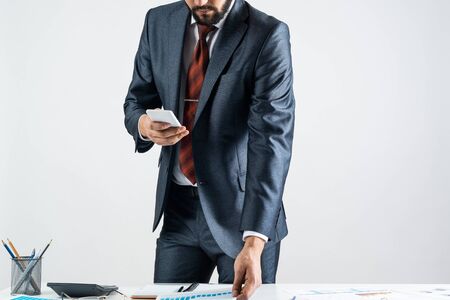 Businessman standing near office desk and using smartphone. Online stock trading and investment. Corporate manager in suit and tie working with business analytics. Entrepreneurship and leadership.の写真素材