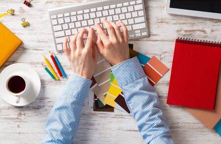 Woman designer typing on keyboard. Creative workspace with color swatches, colorful pencils and cup of coffee. Professional product designing. Coloristics and product branding in visual design studioの写真素材