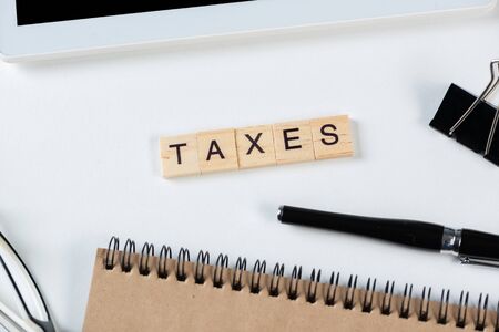 Taxes concept with letters on cubes. Still life of office workplace with supplies. Flat lay white surface with tablet computer, pen and glasses. Company financial management, audit and taxation.の写真素材