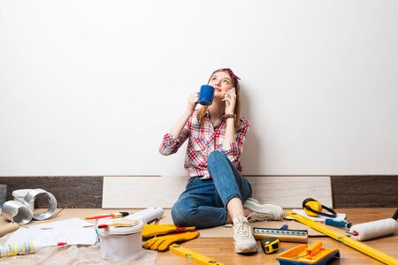 Dreamy girl talking on smartphone at home. Apartment remodeling and house interior renovation concept with copy space. Young attractive woman in red checkered shirt and jeans relaxing with coffee.の写真素材