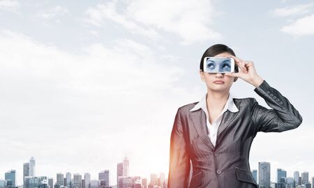 Portrait of pretty woman covering her eyes with smartphone. Businesswoman showing mobile phone with eyes on screen. Corporate businessperson on cityscape background. Mobile communication layout.の写真素材
