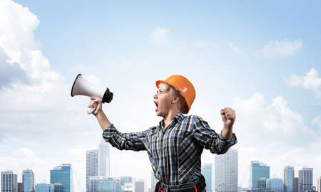 Expressive woman in safety helmet shouting into megaphone. Portrait of young emotional construction worker with wide open mouth on background of modern city. News announcement and advertisement.の写真素材