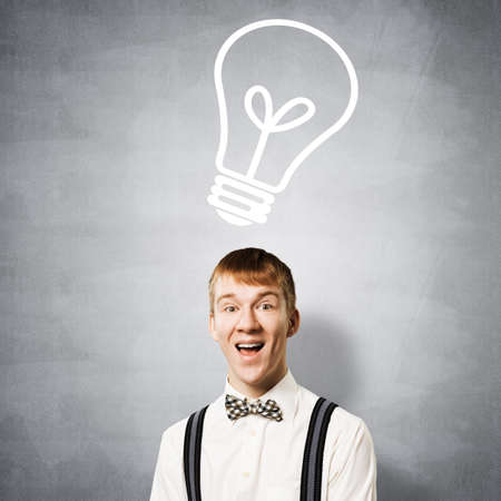 Happy redhead student with broad smile in eureka moment. Creativity and problem solving. Clever boy on background of grey wall with idea lightbulb sketch drawing overhead. Brainstorming conceptの写真素材