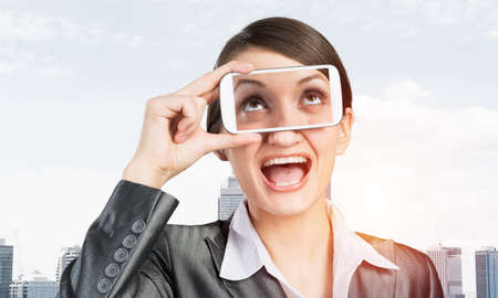 Portrait of pretty woman covering her eyes with smartphone. Happy businesswoman showing mobile phone with eyes on screen. Corporate businessperson on cityscape background. Mobile communication layout.の写真素材