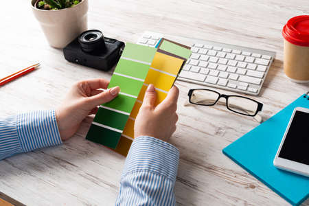 Interior designer holding color swatches at wooden desk. Office workplace with computer keyboard and camera. Brainstorming and creativity. Selection of color palette for web or product design.の写真素材