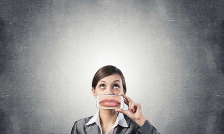 Portrait of young woman covering her mouth with smartphone. Businesswoman showing mobile phone with lips on screen. Corporate businessperson on grey wall background. Mobile communication layout.の写真素材