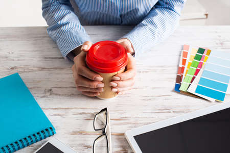 Office manager holding cardboard cup of coffee at wooden desk. Office workspace with tablet computer...