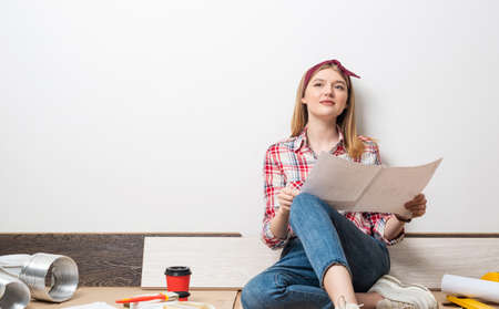 Beautiful girl sitting with paper blueprint in hand. Home remodeling and house interior redesign. Construction tools and materials lying on floor. Young woman studying renovation project of flat.の写真素材