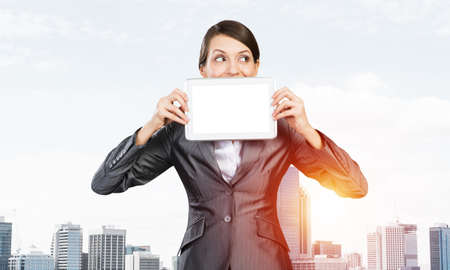 Businesswoman holding tablet computer with blank screen. Beautiful woman in business suit show tablet PC near her face. Corporate businessperson on cityscape background. Digital technology layout.の写真素材