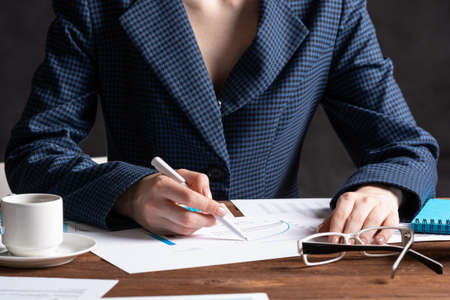 Corporate consultant analyze financial diagram. Close up woman hand with pen and coffee cup on wooden desk. Analyst in suit working at workplace in office. Business accounting and management.の写真素材