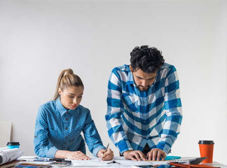 Young man and woman together working at design project. Creative teamwork at workspace with construction blueprint and color swatches. People standing near desk and discussing in architecture studioの写真素材