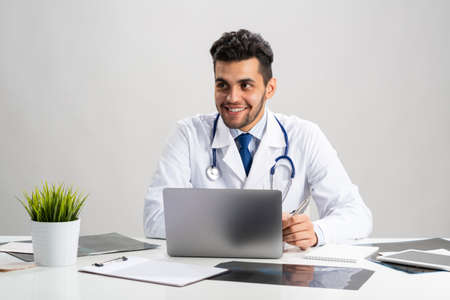 Young arab internist working at computer. Smiling physician in white coat with stethoscope sitting at desk with laptop. Professional medical consultation and treatment in hospital. Medical healthcare.の写真素材