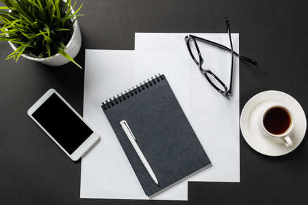 Top view of modern workplace with spiral notepad. Flat lay black surface with smartphone and cup of...