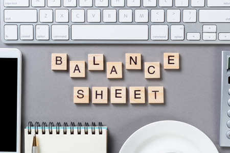 Balance sheet concept with letters on wooden cubes. Still life of office workplace with supplies. Flat lay grey surface with computer keyboard and cup of tea. Budget planning, audit and accounting.の写真素材