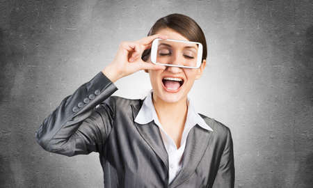 Portrait of pretty woman covering her eyes with smartphone. Happy businesswoman showing mobile phone...