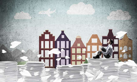 Thoughtful businessman looking away while sitting among flying paper planes with drawn cityscape on...